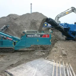 Dwie mobilne kruszarki, Power Screen i Turbo Chieftain 1400, pracują na placu budowy, przesiewając gruz; w tle komin przemysłowy.