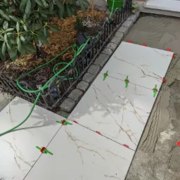 Układanie płytek imitujących marmur na zewnątrz, z użyciem krzyżyków poziomujących, widoczny klej, ogródek z roślinami i zielony wąż ogrodowy w tle.