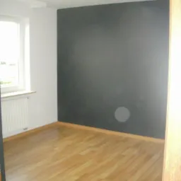 Pokój po renowacji z jasnymi panelami podłogowymi ułożonymi w regularny wzór, ciemną ścianą i białymi ścianami bocznymi z grzejnikiem pod oknem, oświetlony lampą sufitową z kilkoma punktami...