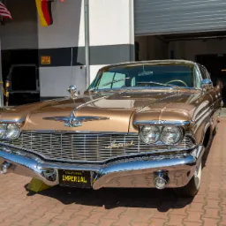 Przód klasycznego, złotego samochodu Imperial z chromowanymi elementami, zaparkowanego przed warsztatem z otwartą bramą garażową i wiszącymi flagami.