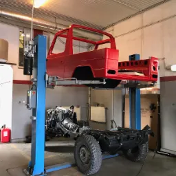 Czerwona karoseria samochodu podniesiona na podnośniku warsztatowym, pod nią podwozie z silnikiem, widoczny fragment warsztatu z oświetleniem i elementami wyposażenia.