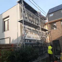 Ocieplanie elewacji domu jednorodzinnego z użyciem rusztowania; widoczna siatka z fragmentami ocieplenia i pracownik w żółtej kamizelce sprzątający teren wokół budynku.