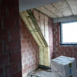 Wnętrze w trakcie prac związanych z izolacją termiczną poddasza: widoczne wełna mineralna między krokwiami, fragment ściany z czerwonej cegły oraz opakowanie styropianu EPS na podłodze.