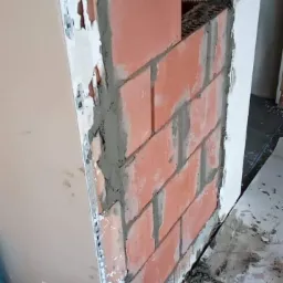 Wznoszenie ścianki z cegły klinkierowej wokół komina; widoczny fragment instalacji kominowej, zaprawa murarska i listwa narożna.