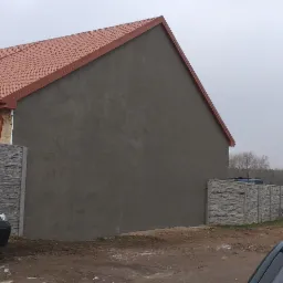 Szara elewacja budynku z czerwoną dachówką, widoczna zza betonowego ogrodzenia w pochmurny dzień. Fragment elewacji w trakcie wykonywania, widoczna surowa ściana oraz fragment starej elewacji...