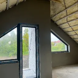 Poddasze w trakcie wykańczania, ściany po tynkowaniu maszynowym, widoczne okna dachowe i izolacja pianą PUR na poddaszu, profile okienne z logo VEKA.