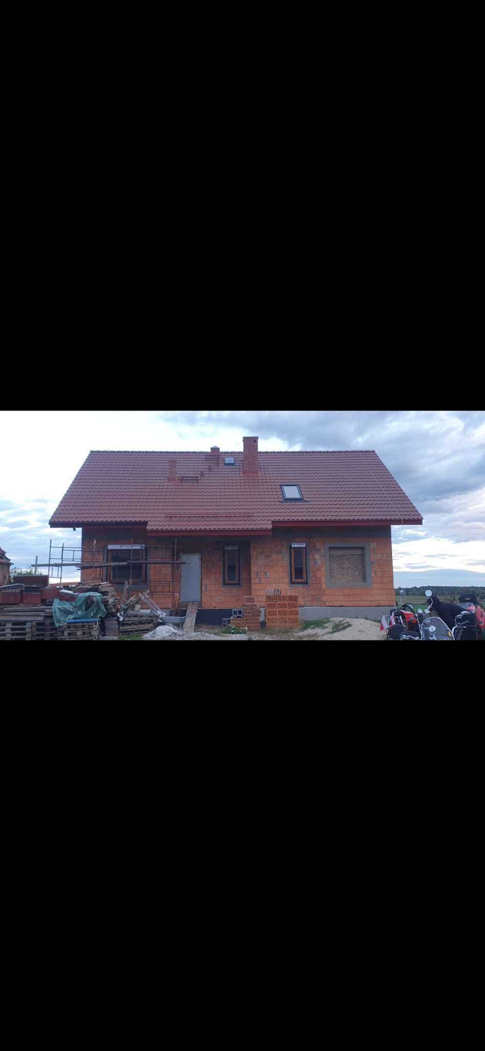 Niedokończony dom z cegły z czerwoną dachówką w trakcie budowy. Widoczne rusztowania, okna i drzwi. Częściowo zachmurzone niebo w tle.