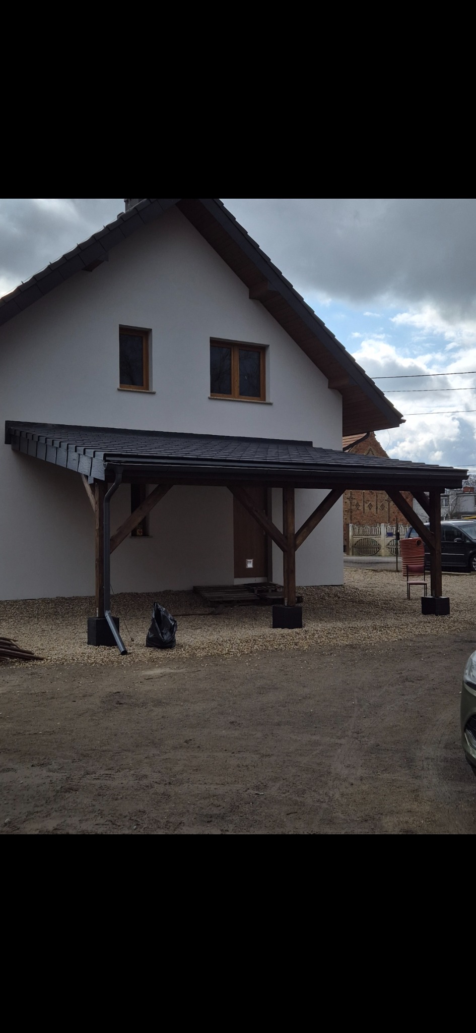 Zadaszenie nad wejściem do domu z czarną dachówką i drewnianą konstrukcją. Nowoczesny design, jasna elewacja. Widok z zewnątrz, dzień.