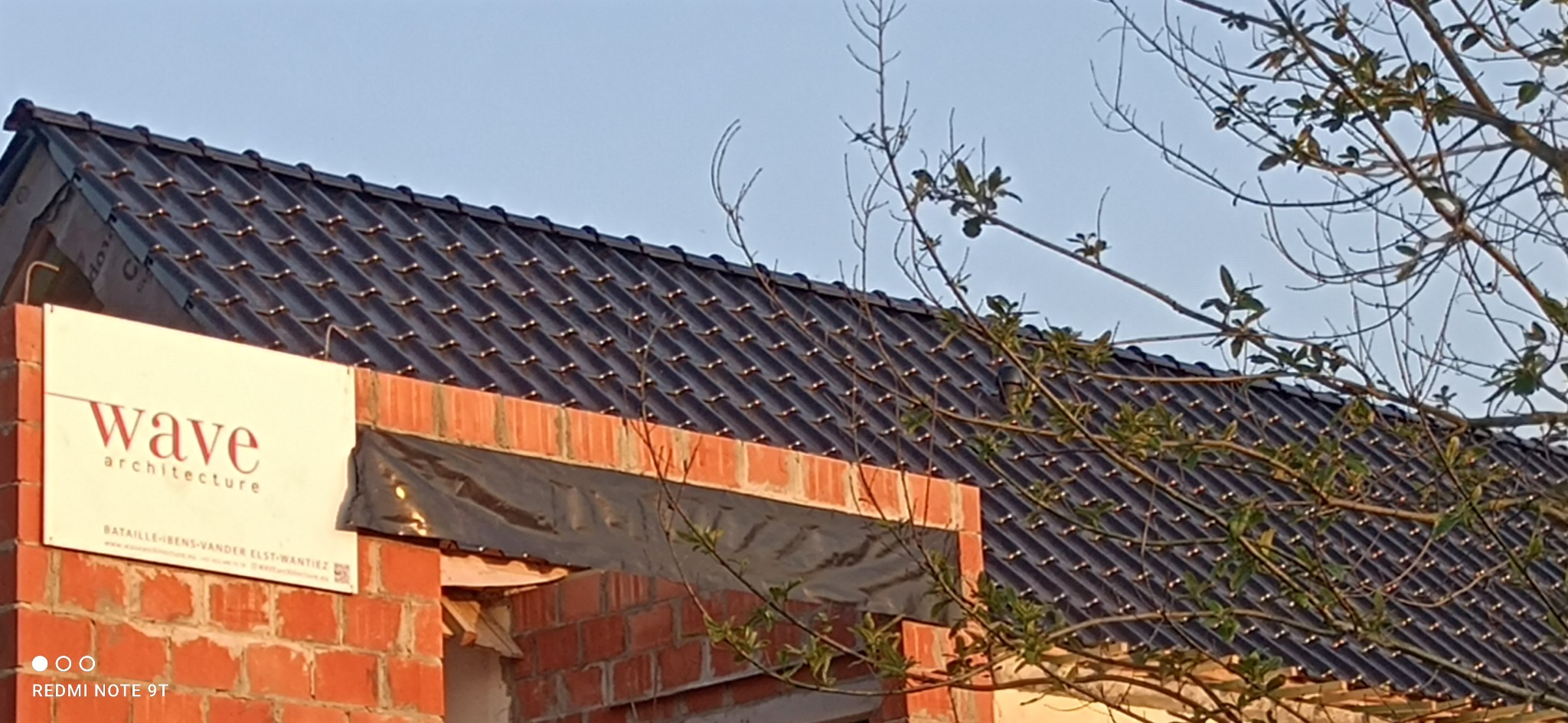 Fragment dachu z czarnej dachówki, widoczny spod gałęzi drzewa, z ceglaną ścianą w stanie surowym i tablicą reklamową firmy architektonicznej.