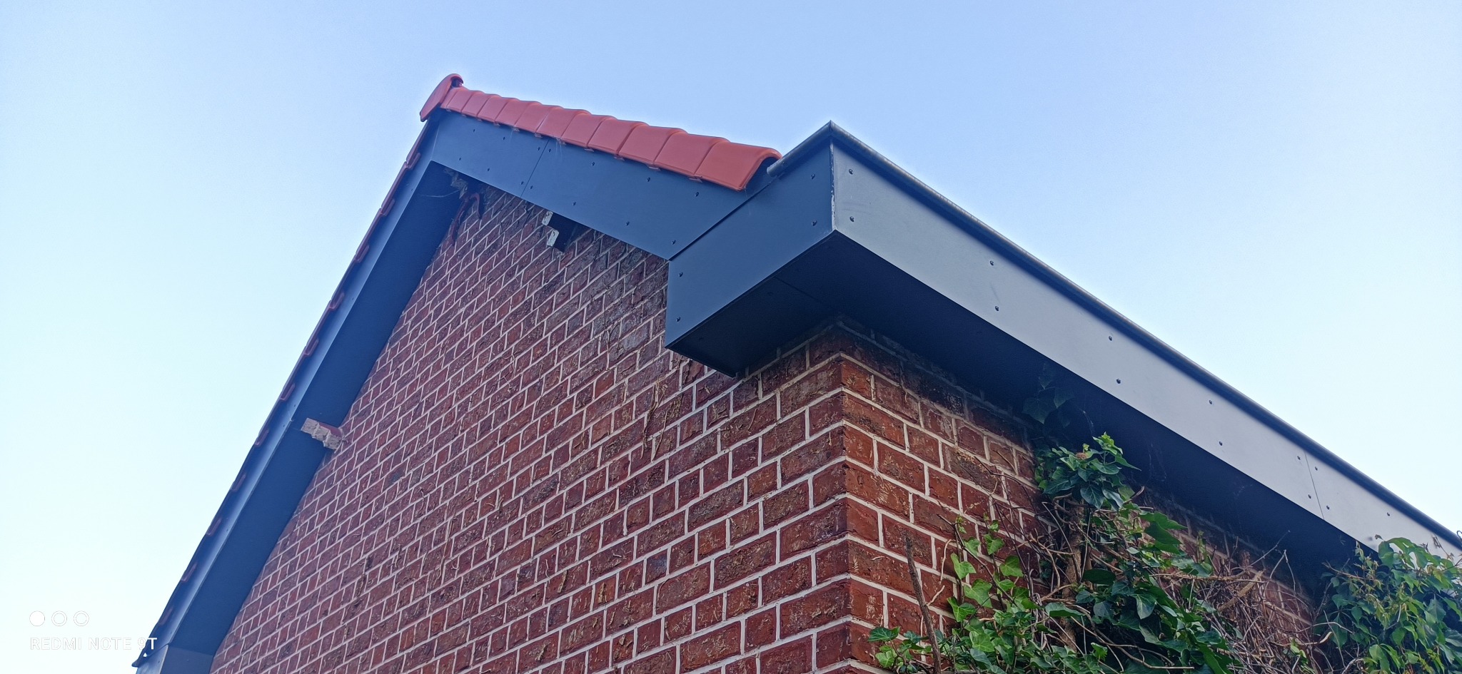 Fragment narożnika domu z czerwonej cegły z nowym, szarym, metalowym orynnowaniem i fragmentem dachu pokrytego czerwoną dachówką. Na ścianie widoczne pnącze.