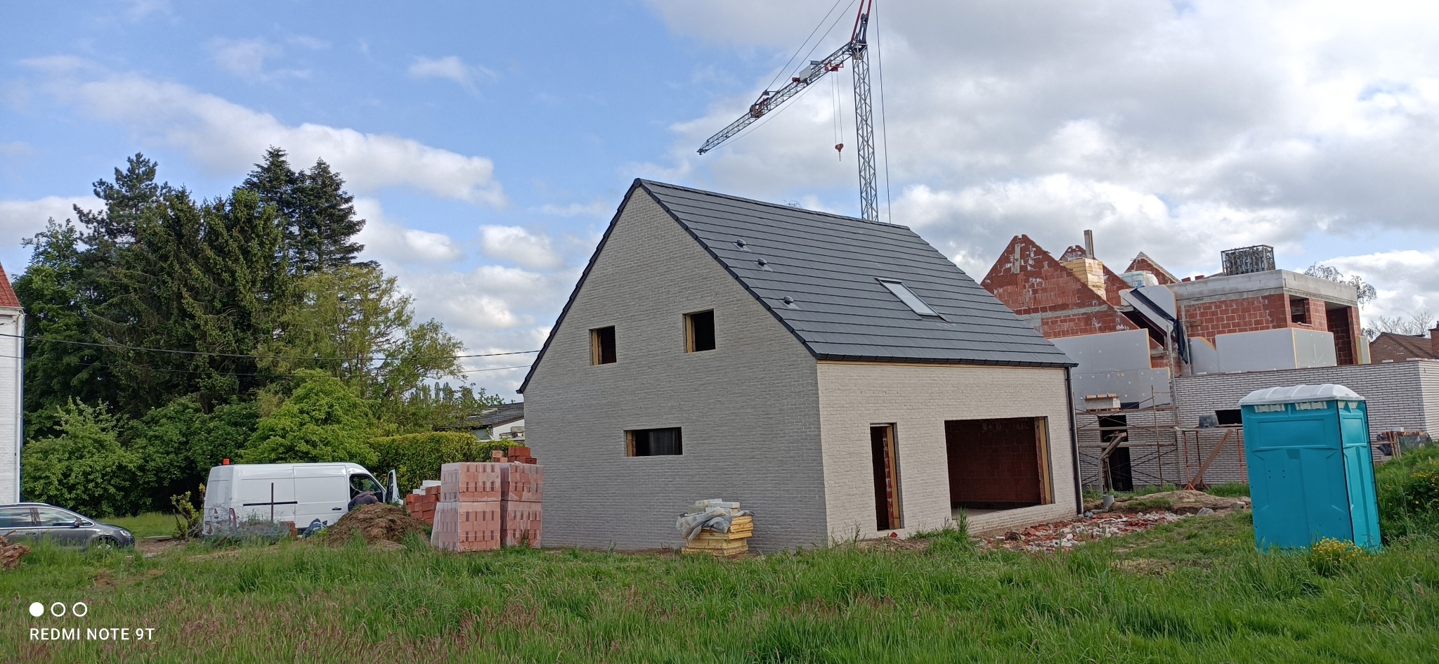 Nowo wybudowany dom z szarym dachem i oknami bez szyb, widoczny dźwig budowlany w tle, sterta cegieł i przenośna toaleta na trawniku.