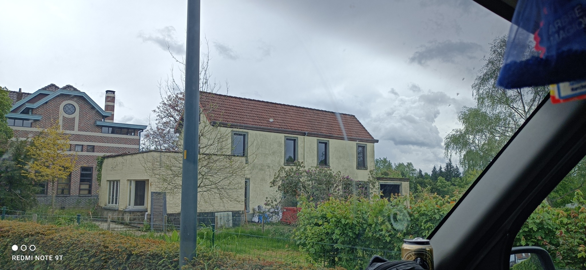 Dom z czerwoną dachówką widziany z wnętrza pojazdu, z widocznymi elementami deski rozdzielczej i zawieszką zapachową, w tle pochmurne niebo.