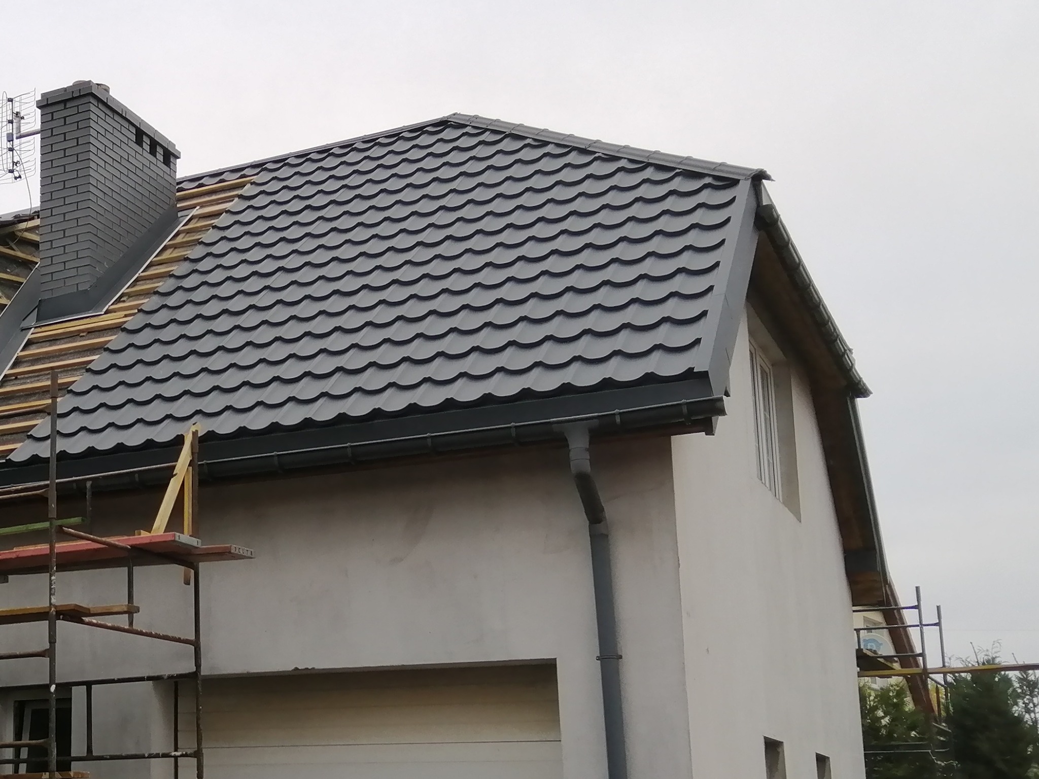 Dom w trakcie prac dekarskich: nowa, grafitowa blachodachówka ułożona na części dachu, widoczne elementy drewnianej konstrukcji, komin z szarej cegły, rusztowanie przy ścianie budynku.