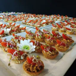 Tarty z nadzieniem, pomidorem i kiełkami, ułożone na białych tacach, z dekoracyjnym białym kwiatem pośród nich, na tle czarnej ściany.