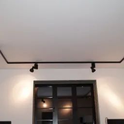 Czarna szyna oświetleniowa z reflektorami punktowymi zamontowana na suficie w jasnym pomieszczeniu z marmurową ścianą i oknem z ciemną ramą.