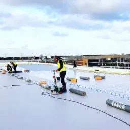 Pracownicy w kamizelkach odblaskowych wykonują prace na płaskim dachu, używając zgrzewarki do łączenia rolek materiału hydroizolacyjnego.