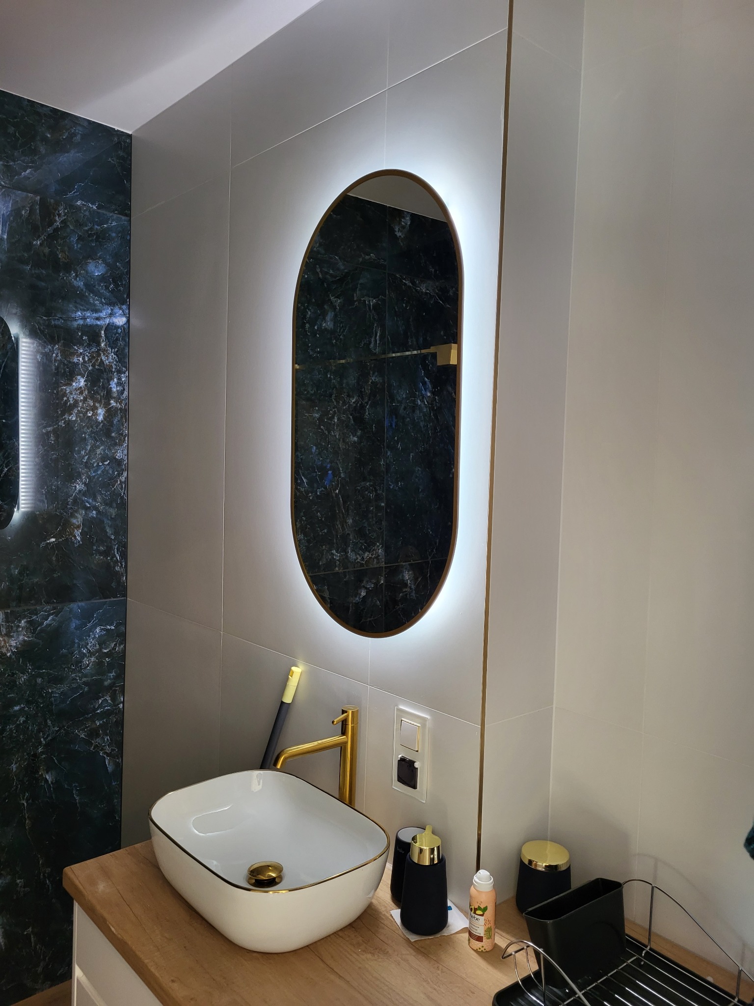 Nowoczesna łazienka z owalnym lustrem podświetlanym LED, białym blatem i umywalką nablatową z baterią w kolorze złota. Ściany wyłożone płytkami imitującymi marmur.