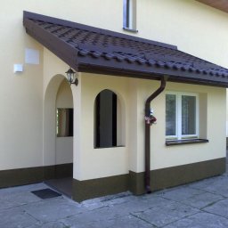 MICHAŁ JAROS USŁUGI REMONTOWO-BUDOWLANE, INSTALACJE SANITARNE REMONTIS - Otynkowana elewacja domu z gankiem, brązowy dach i rynna, widoczne okno z kwiatami i lampa zewnętrzna.