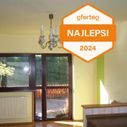 Wnętrze pokoju po odświeżeniu, z jasnymi ścianami w odcieniach żółci i zieleni, widoczne okno balkonowe z brązową ramą, grzejnik oraz fragment kuchni z kafelkami. Na zdjęciu naklejka Oferteo...