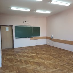 MICHAŁ JAROS USŁUGI REMONTOWO-BUDOWLANE, INSTALACJE SANITARNE REMONTIS - Wnętrze klasy po remoncie: parkiet w jodełkę, ściany w odcieniach różu, tablica i listwy przypodłogowe. Widoczne lampy sufitowe i gniazdka elektryczne.