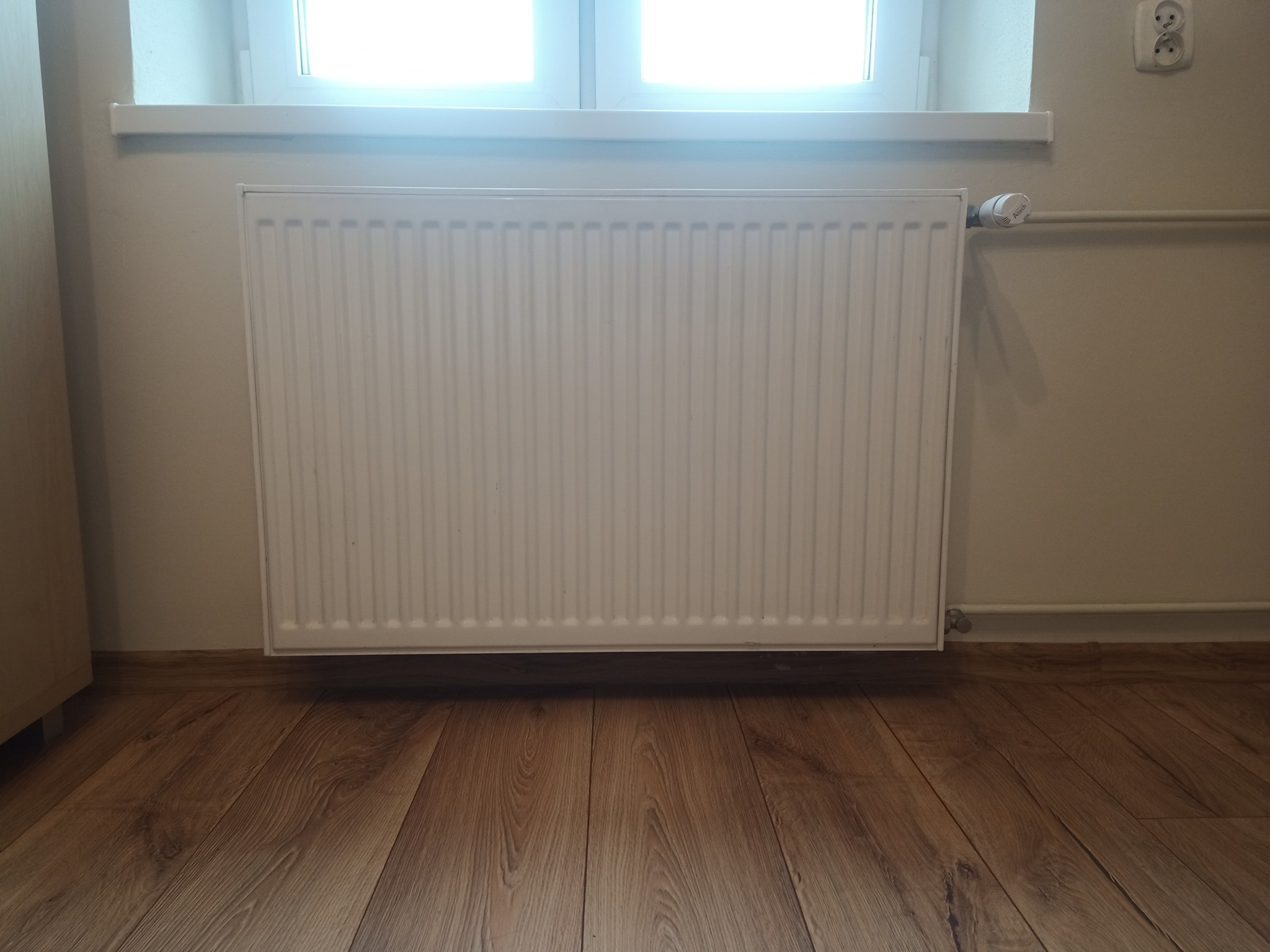 Biały, panelowy grzejnik zamontowany pod oknem na tle drewnopodobnej podłogi i beżowej ściany z widocznym gniazdkiem elektrycznym.