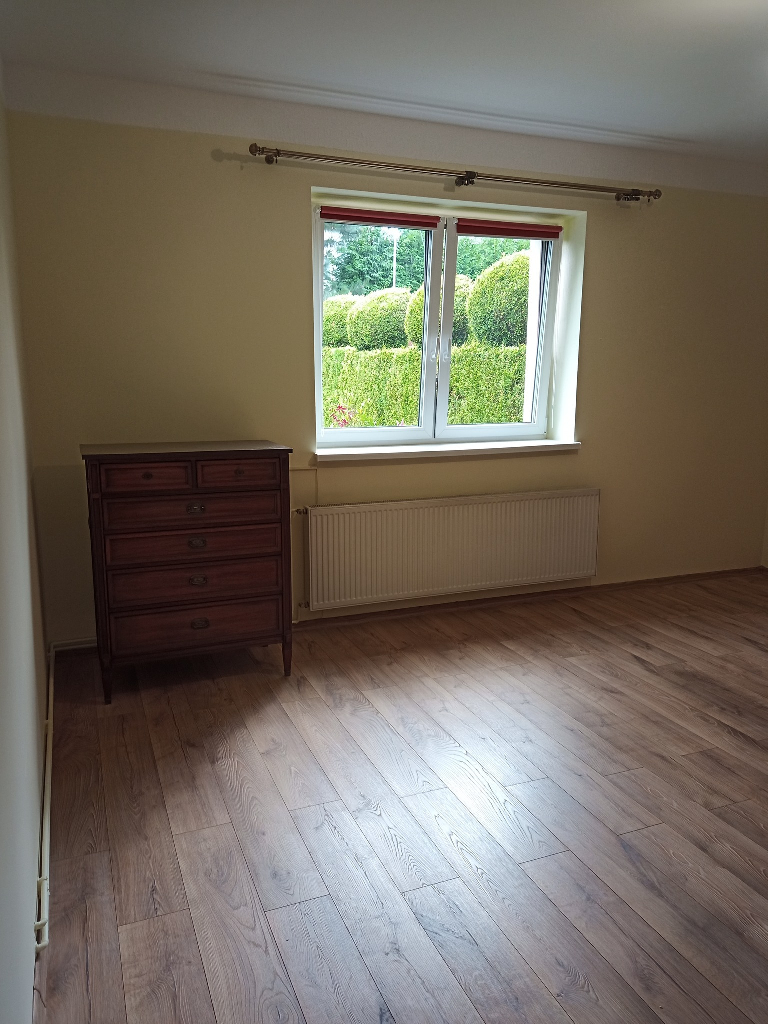 Pokój z nowo ułożonymi panelami podłogowymi w odcieniach brązu, widok na okno z zielonym żywopłotem i komodę z ciemnego drewna