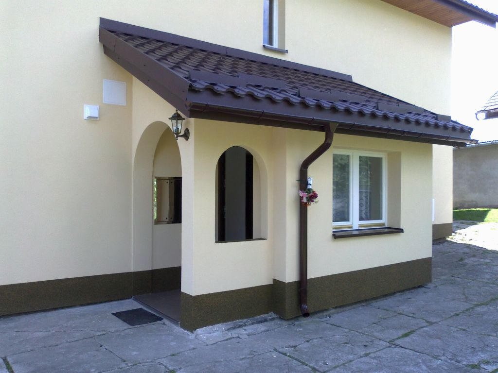Otynkowana elewacja domu z gankiem, brązowy dach i rynna, widoczne okno z kwiatami i lampa zewnętrzna.