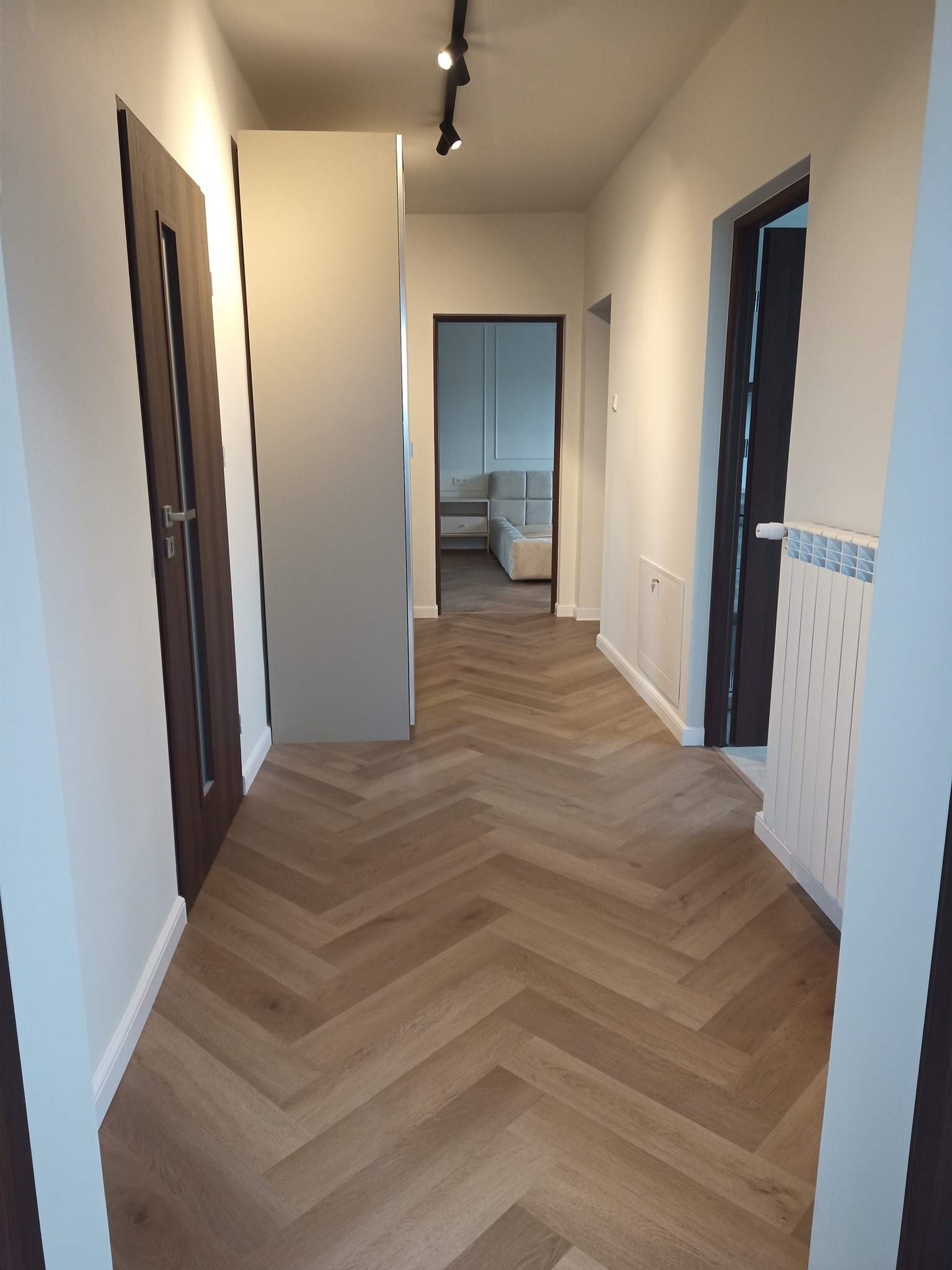 Korytarz z podłogą ułożoną w jodełkę z paneli, widoczne drzwi wewnętrzne z ciemnego drewna, białe ściany i grzejnik żeberkowy, w tle widoczny fragment sypialni z łóżkiem.