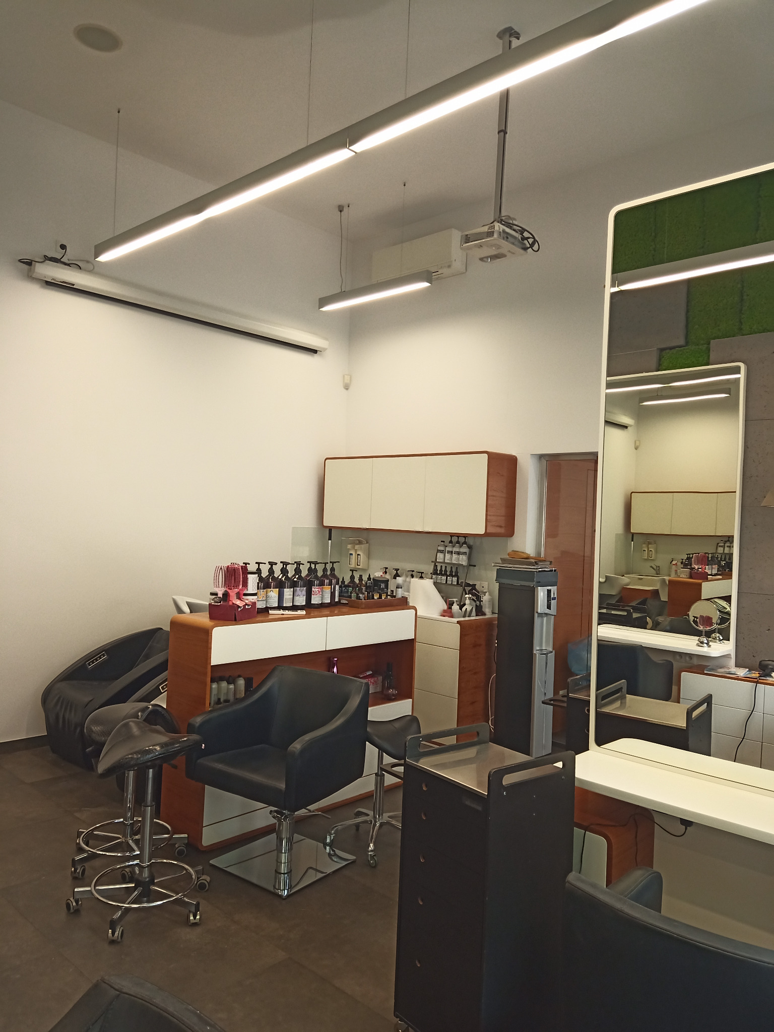 Nowoczesne wnętrze salonu fryzjerskiego z czarnymi fotelami, lustrami i oświetleniem liniowym. Widoczne stanowiska pracy i produkty do pielęgnacji włosów.
