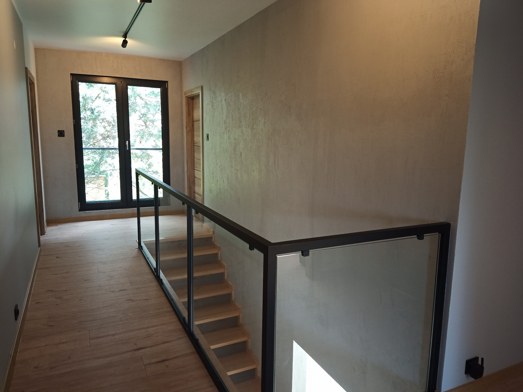 Korytarz z drewnianą podłogą, schodami z balustradą ze szkła i czarnych ram. Ściany w odcieniach beżu, okno z widokiem na zieleń. Nowoczesne oświetlenie.