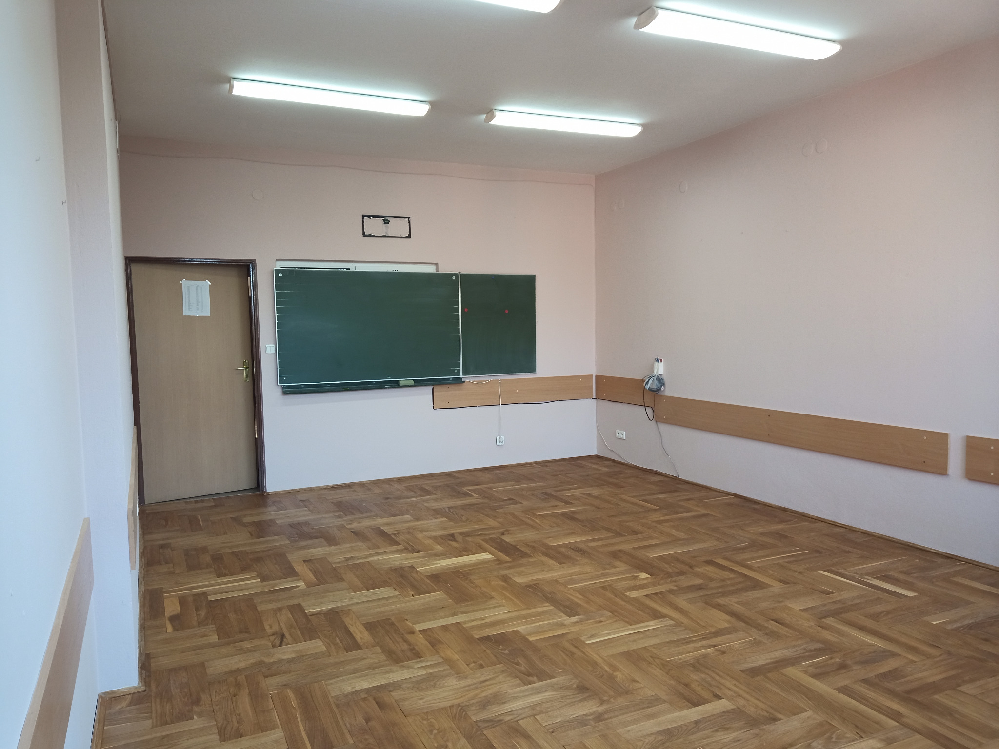 Wnętrze klasy po remoncie: parkiet w jodełkę, ściany w odcieniach różu, tablica i listwy przypodłogowe. Widoczne lampy sufitowe i gniazdka elektryczne.