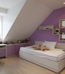 Pokój dziecięcy na poddaszu z fioletową ścianą, białym łóżkiem i zabudową meblową z fioletowymi frontami. Na łóżku leży pluszowy miś. Widoczne skosy dachu i białe, drewniane podłogi.