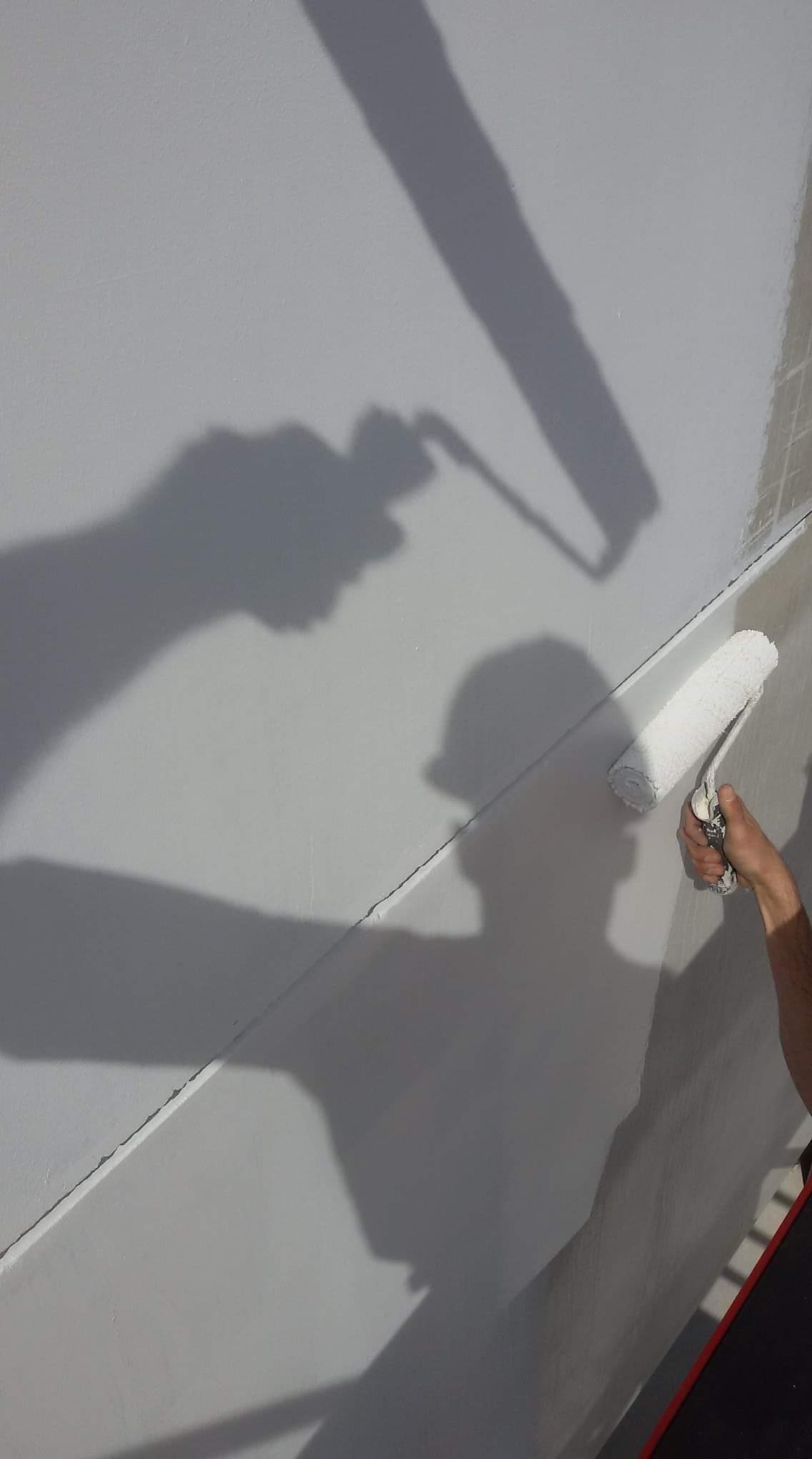Cień malarza z wałkiem na jasnej ścianie podczas malowania, widoczna faktura świeżej farby i fragment drabiny.