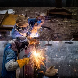 Dwóch spawaczy łączy metalowe elementy, iskry rozświetlają ciemne otoczenie, widoczne ochronne maski i rękawice.