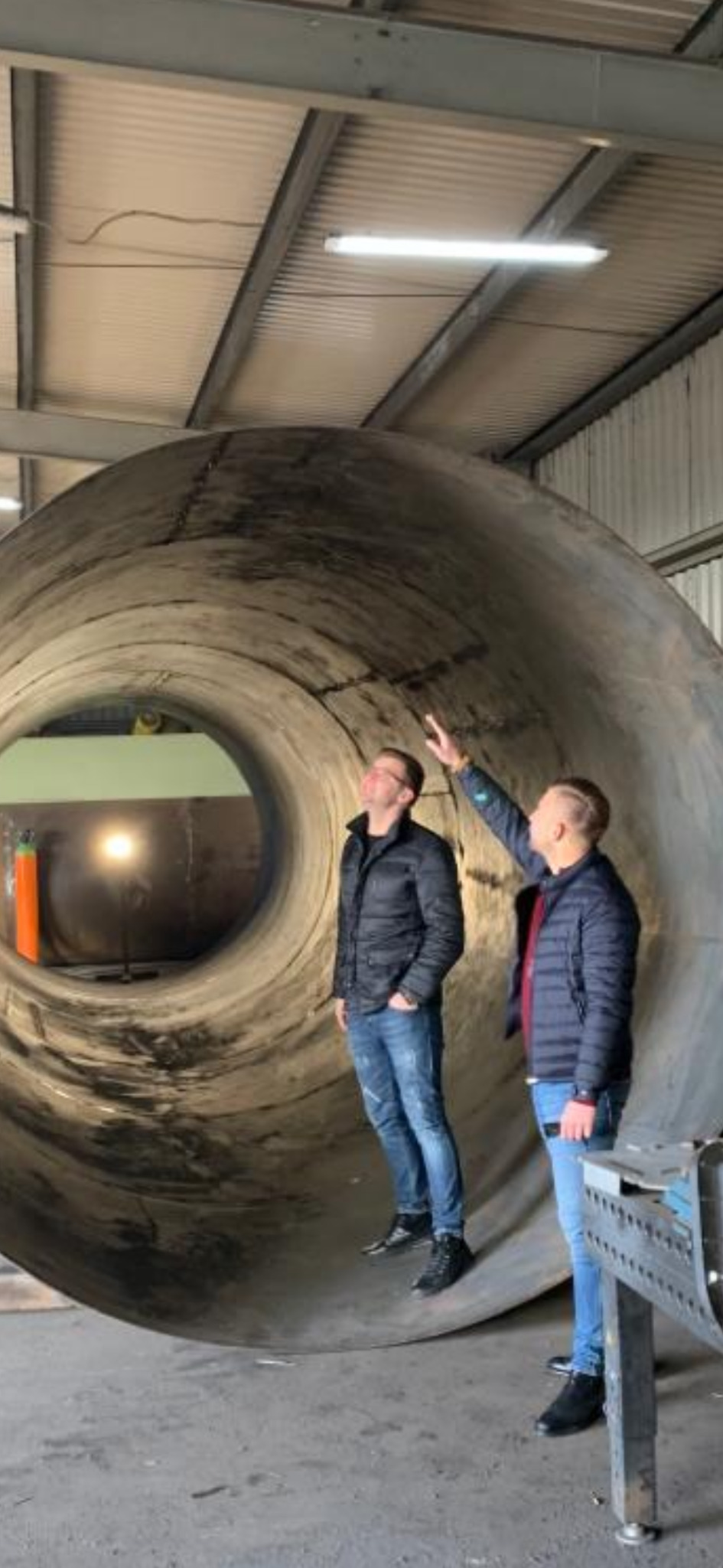 Dwaj mężczyźni w kurtkach i jeansach stoją przed ogromną, spawaną rurą z metalu, jeden wskazuje palcem na górę konstrukcji, w tle widoczny warsztat z oświetleniem.
