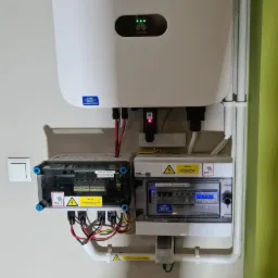 Biały inwerter marki Huawei z trzema świecącymi diodami, zamontowany na ścianie nad dwoma skrzynkami rozdzielczymi z widocznymi bezpiecznikami i okablowaniem, w tle fragment zielonej ściany.