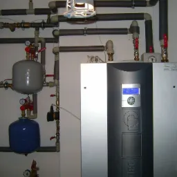 Instalacja grzewcza z szarym zbiornikiem wyrównawczym i niebieskim naczyniem przeponowym, liczne rury z izolacją, panel sterowania z wyświetlaczem LCD, widok z przodu.