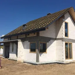 Surowy budynek jednorodzinny w trakcie budowy, widoczne ściany z betonu komórkowego, częściowo otynkowane, z oknami w drewnianych ramach, dach z deskowaniem pod pokrycie dachowe, paleta z cegłami...