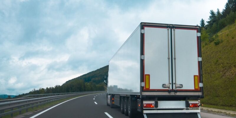 Biała naczepa ciężarówki w ruchu na autostradzie, widok z tyłu, krajobraz zalesionych wzgórz w tle pod pochmurnym niebem.