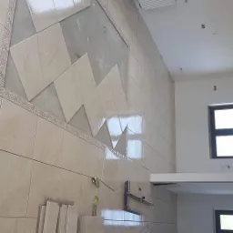 Układanie beżowych płytek na ścianie z dekoracyjnym pasem, widoczny grzejnik, okno i narzędzia glazurnicze.