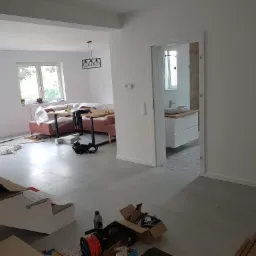 Wnętrze w trakcie remontu: widok na salon z różową sofą i jadalnią, otwartą łazienkę z umywalką na szafce oraz prowizoryczne schody zabezpieczone kartonem i taśmą malarską, narzędzia i materiały...