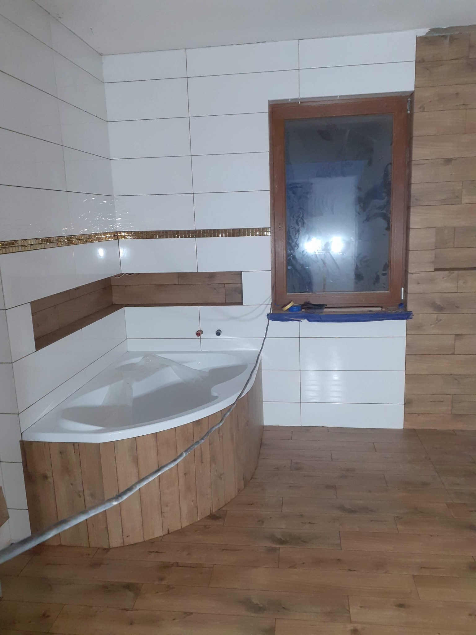 Narożna wanna obudowana drewnopodobnymi panelami, białe płytki ceramiczne z dekoracyjnym pasem, drewnopodobne panele na ścianie obok okna, podłoga z drewnopodobnych paneli.