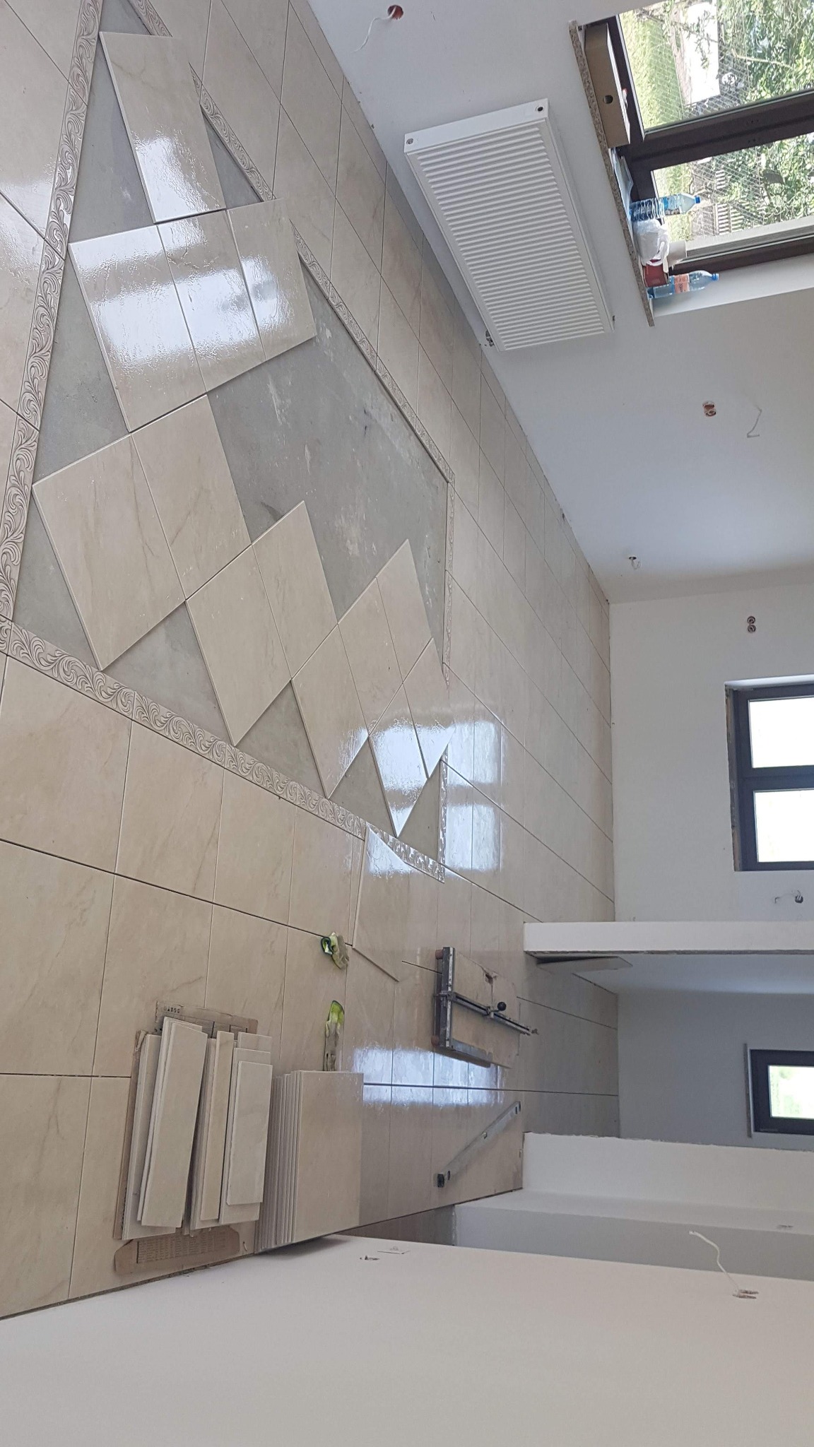 Układanie beżowych płytek na ścianie z dekoracyjnym pasem, widoczny grzejnik, okno i narzędzia glazurnicze.