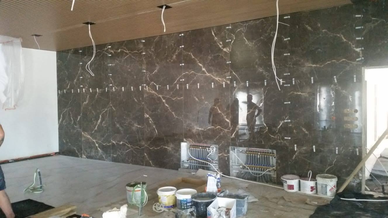 Montaż ciemnych, marmurkowych płytek wielkoformatowych na ścianie, widoczne plastikowe krzyżyki dystansowe, instalacja ogrzewania podłogowego w ścianie, puszki z kablami wystającymi z sufitu...