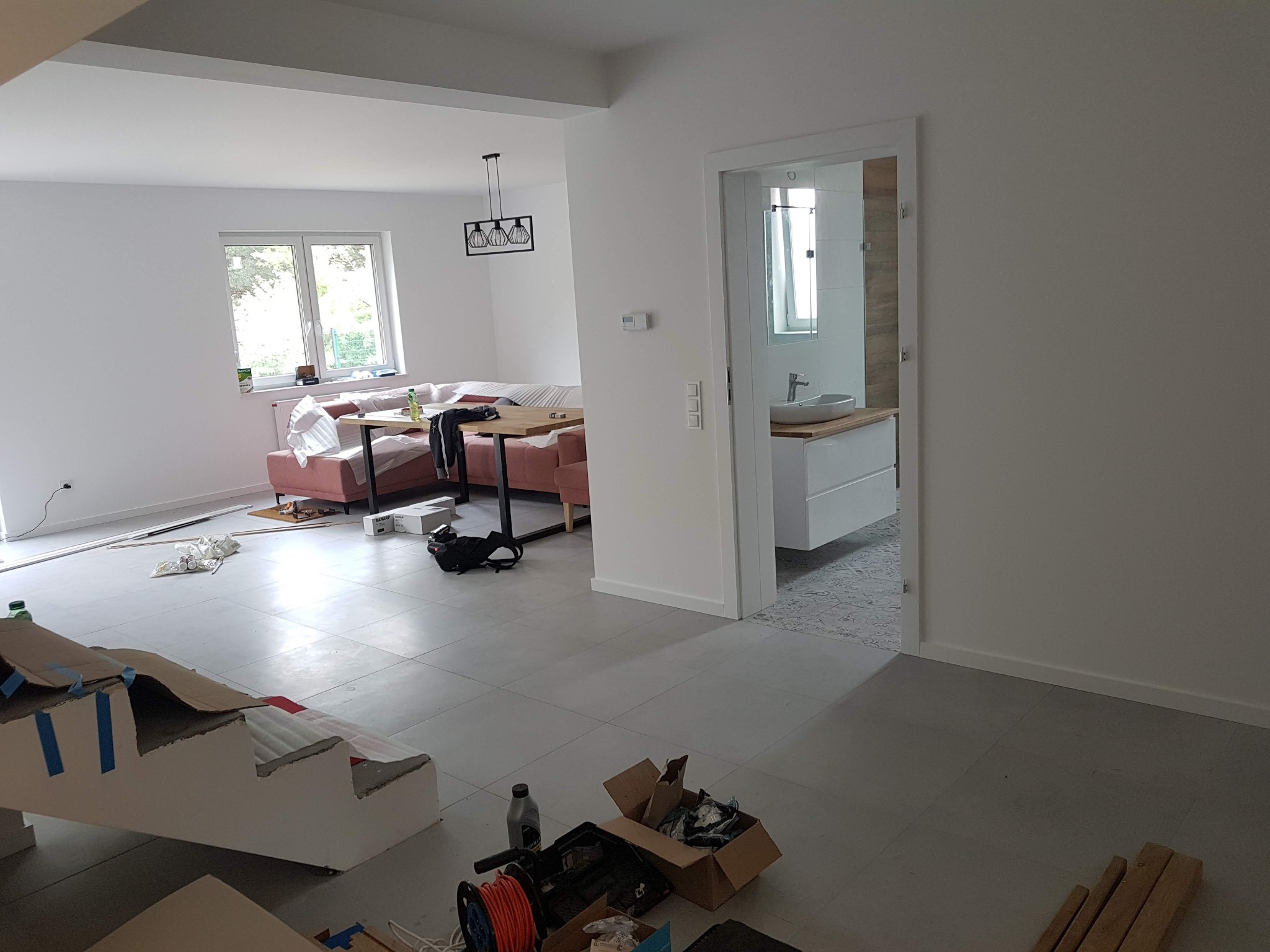 Wnętrze w trakcie remontu: widok na salon z różową sofą i jadalnią, otwartą łazienkę z umywalką na szafce oraz prowizoryczne schody zabezpieczone kartonem i taśmą malarską, narzędzia i materiały...