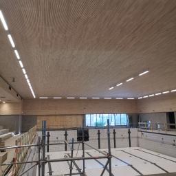 Alex-Bedrijf - Montaż sufitu z drewnianych paneli w hali basenowej, widoczne rusztowania, oświetlenie liniowe LED i niecka basenowa z białymi płytkami.