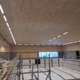 Montaż sufitu z drewnianych paneli w hali basenowej, widoczne rusztowania, oświetlenie liniowe LED i niecka basenowa z białymi płytkami.