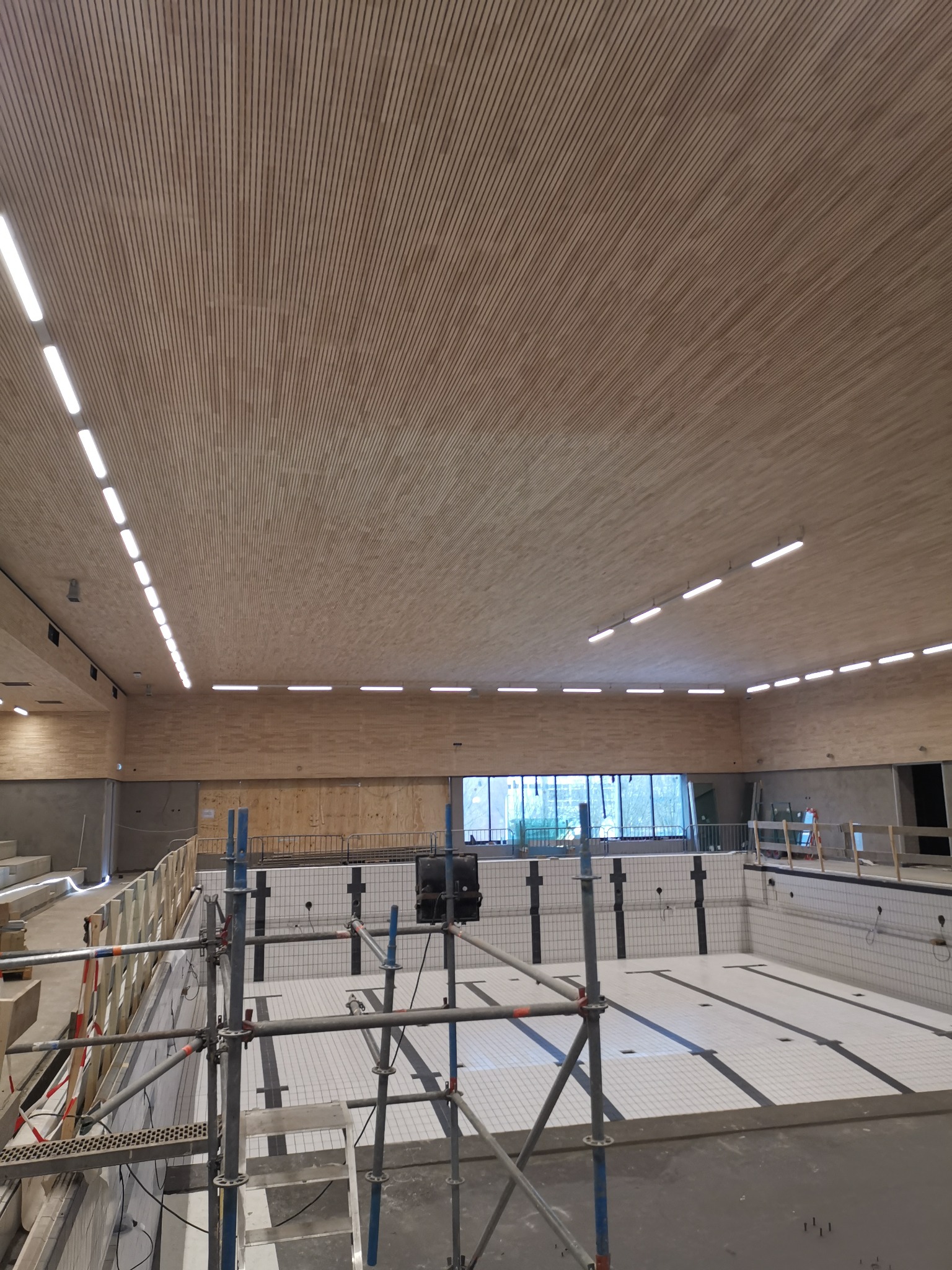 Montaż sufitu z drewnianych paneli w hali basenowej, widoczne rusztowania, oświetlenie liniowe LED i niecka basenowa z białymi płytkami.