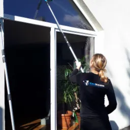 Pracownica firmy sprzątającej EmiHouse czyści wysokie okno za pomocą mopa teleskopowego z niebieską nakładką, widoczny budynek z białą elewacją i kostka brukowa.