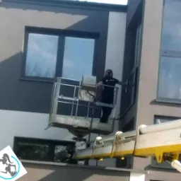 Pracownik w podnośniku koszowym przy budynku z szarymi i białymi panelami, myjący okna na wysokości. Widoczne odbicie nieba w oknach.